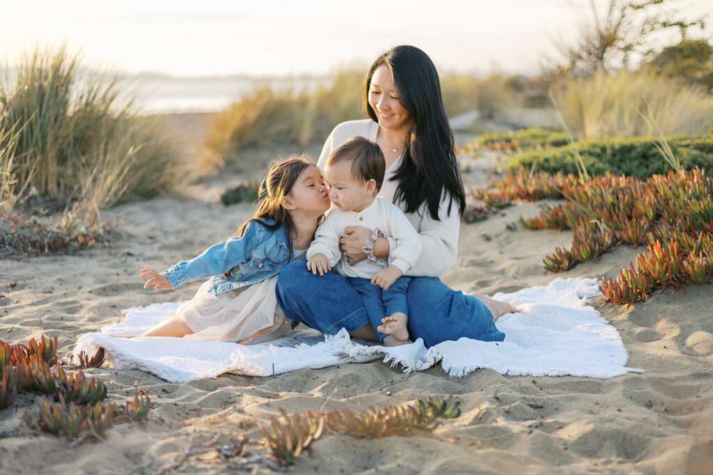 Mom sits on a blue blanket at Alameda Beach with two small children. Older sister leans over to kiss baby brother during a Bay Area family photography session