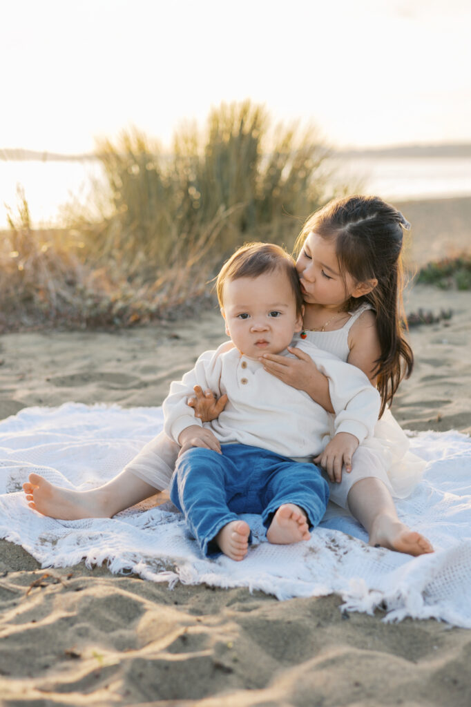 sister kisses baby brother while sitting on alameda beach during East Bay family photography session