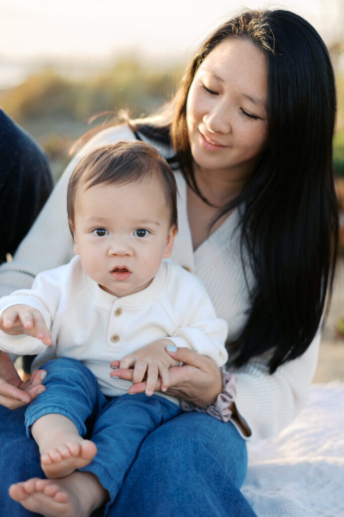 mom leans over baby boy while sitting on Alameda Beach during Bay Area family photography session