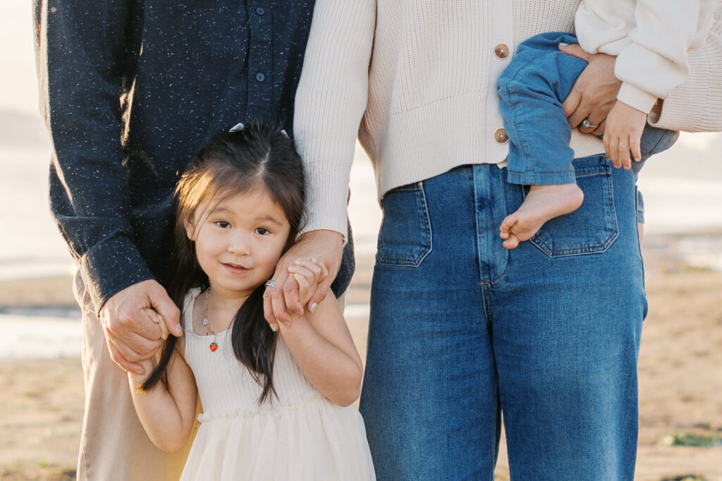toddler girl looks at the camera while holding her parents hands on Alameda Beach during a East Bay family photography session