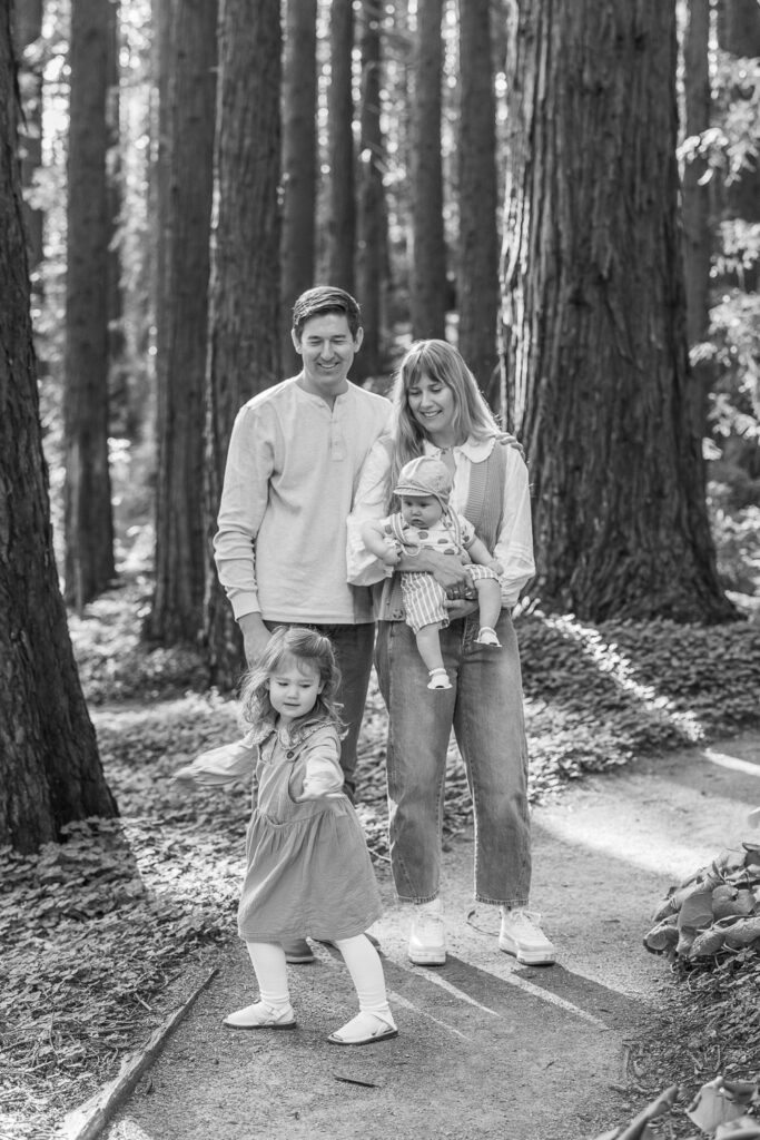 family of four pose in the redwood grove while toddler twirls in front of them during Berkeley family photography session