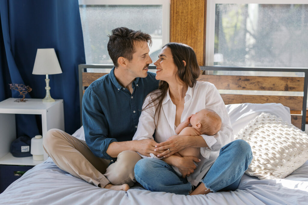 new parents lean in for a kiss while holding 2 month old baby boy new parents hold newborn while sitting on bed with dog for San Francisco newborn photography session