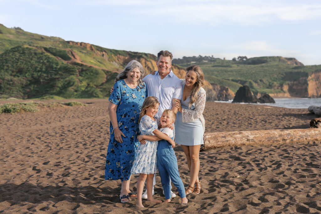 family of four plus grandma pose for a photo on rodeo beach in marin county during bay area family photography session
