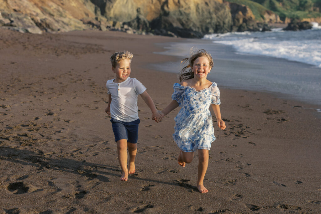 two young siblings run hand in hand on rodeo beach