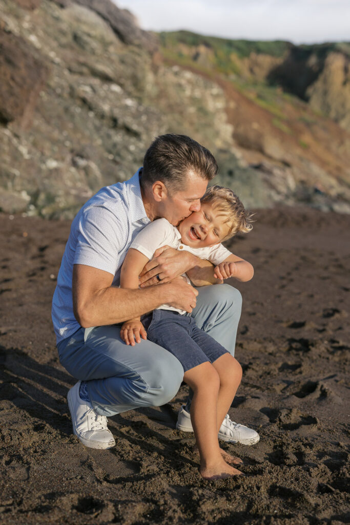 dad leans in and kisses young boy's cheek during family photography session on bay area beach rodeo beach in marin