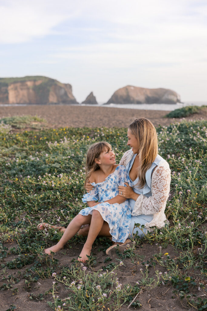 mother and daughter sit in wildflowers on rodeo beach, laughing during bay area family photography session