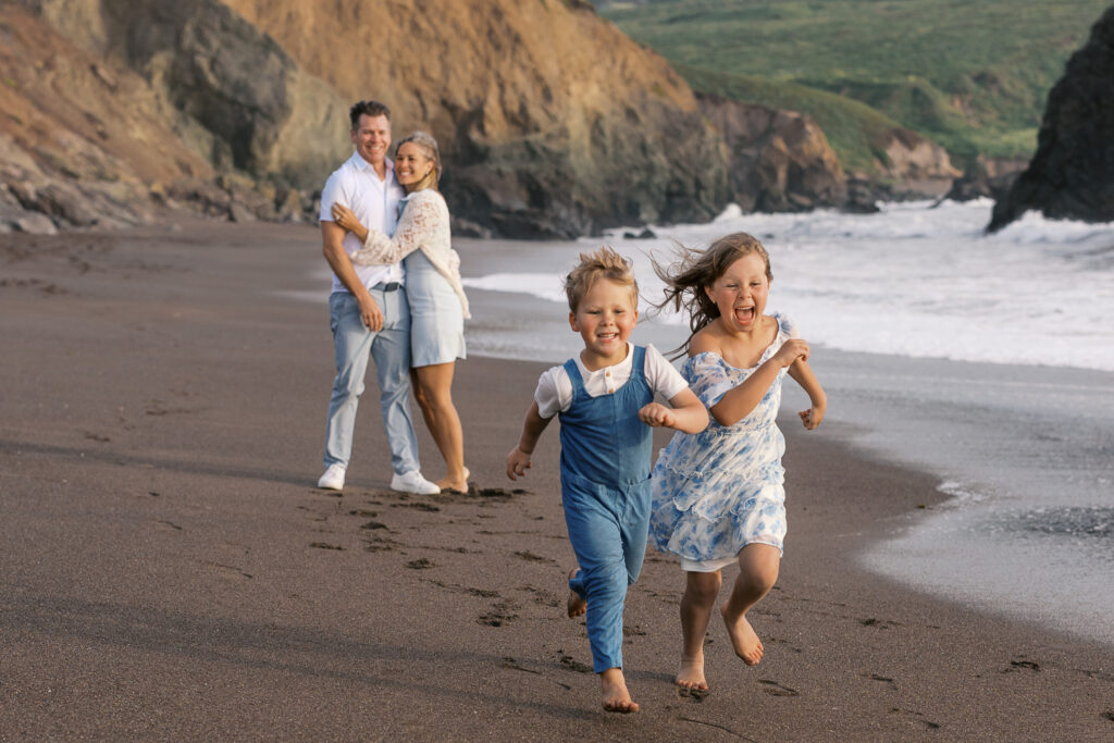 family of four on rodeo beach, mom and dad snuggling while two young children run free