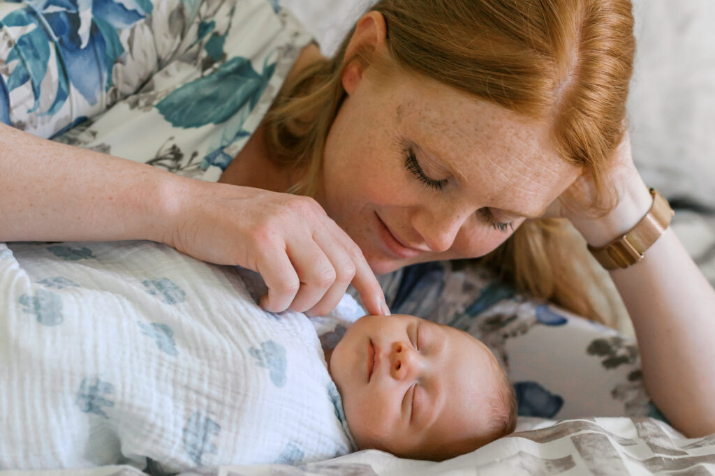 mom lays on bed with newborn baby girl and gently touches her cheek during Alameda in home newborn session