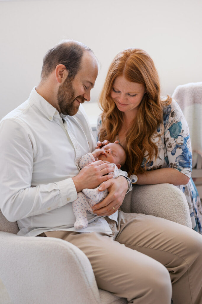parents gaze down at their newborn whiele sitting in a chair during in home photography session