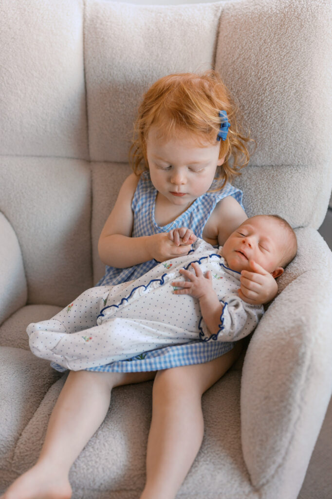 toddler big sister holds newborn baby sister and looks at her tiny hands during an in home newborn session in Alameda