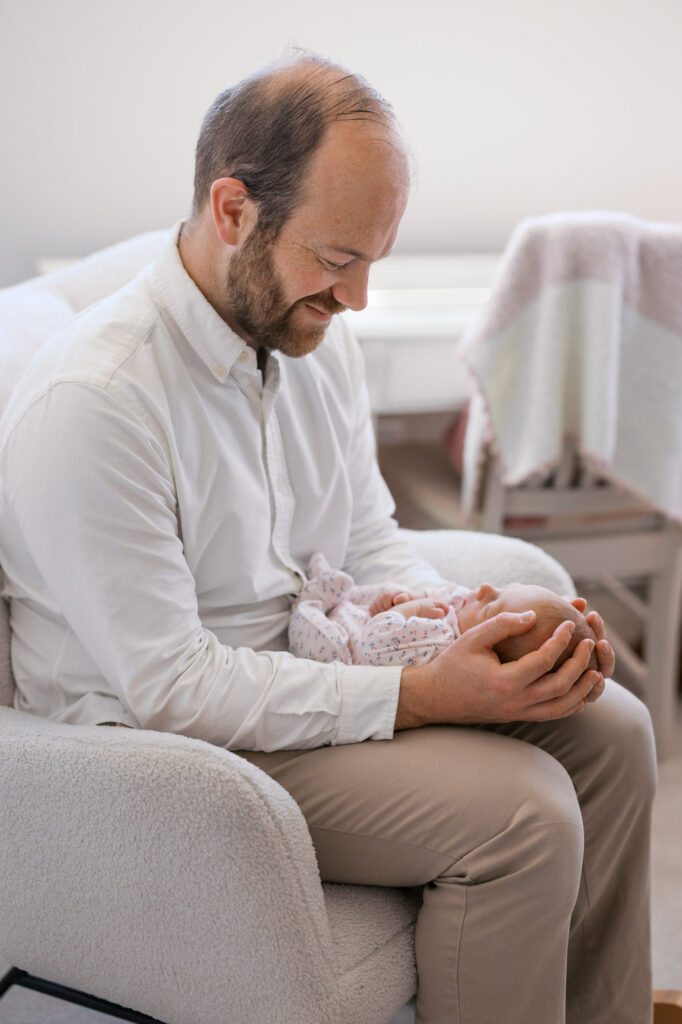 dad holds newborn baby girl and gazes down at how tiny she is in his arms during newborn session in Alameda