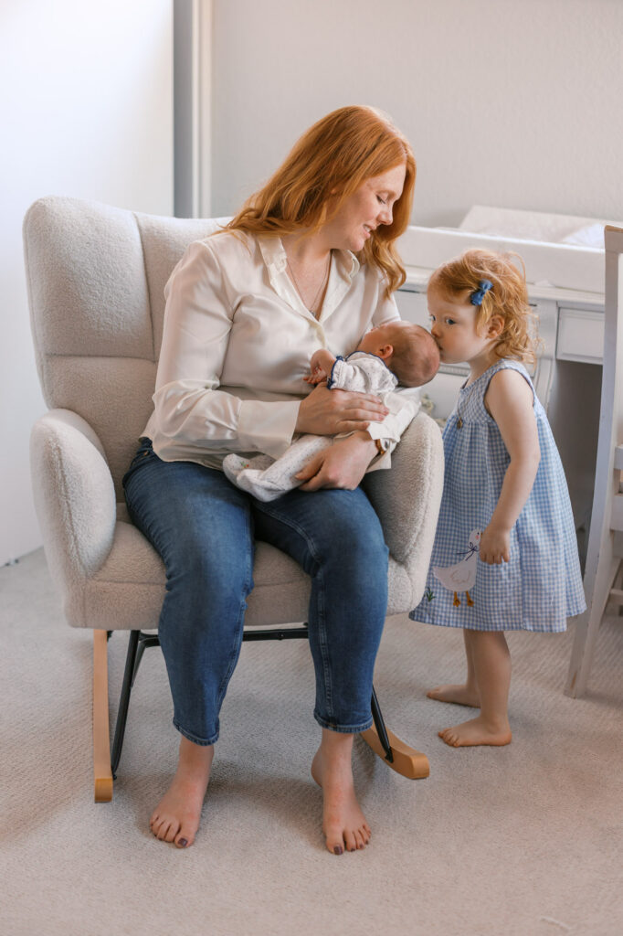 mom holds newborn baby girl while big sister comes over to give her a kiss during in home newborn session