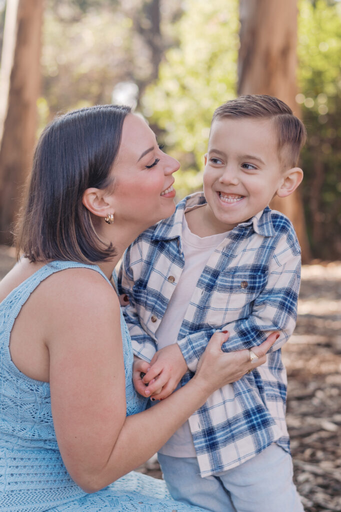 Mom hugs young boy who laughs in eucalyptus grove during a Berkeley family photography session on UC Berkeley campus