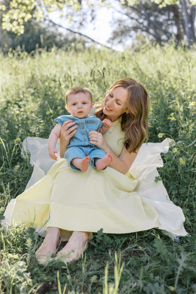 Mom wearing a yellow dress gazes at baby boy in blue romper while sitting on grass with yellow flowers in the Oakland Hills during an East Bay family photography session