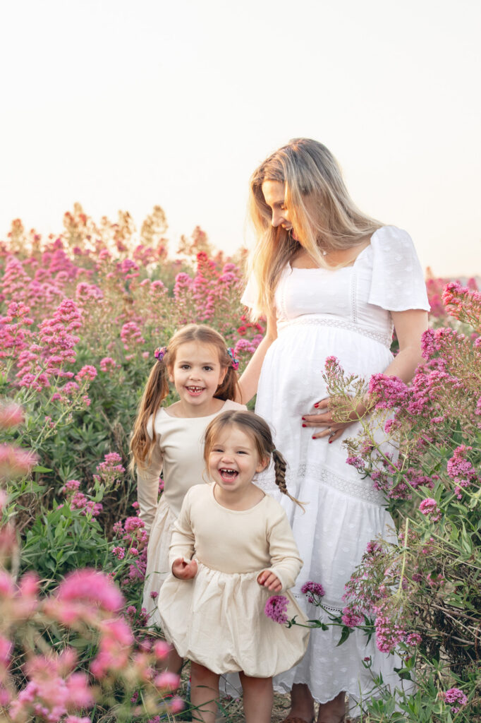 Expecting mom with her two young daughters play in a pink flower field in East Bay during Bay Area maternity photography session