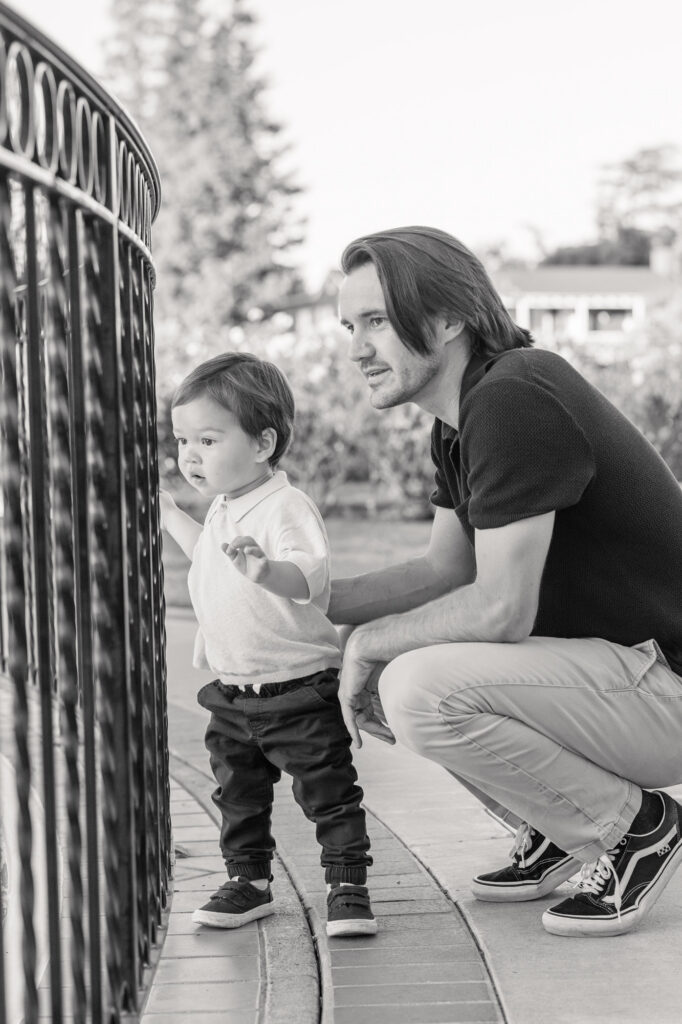 Father crouches next to son who is watching the water fountain at San Jose Rose Garden during a family photography session