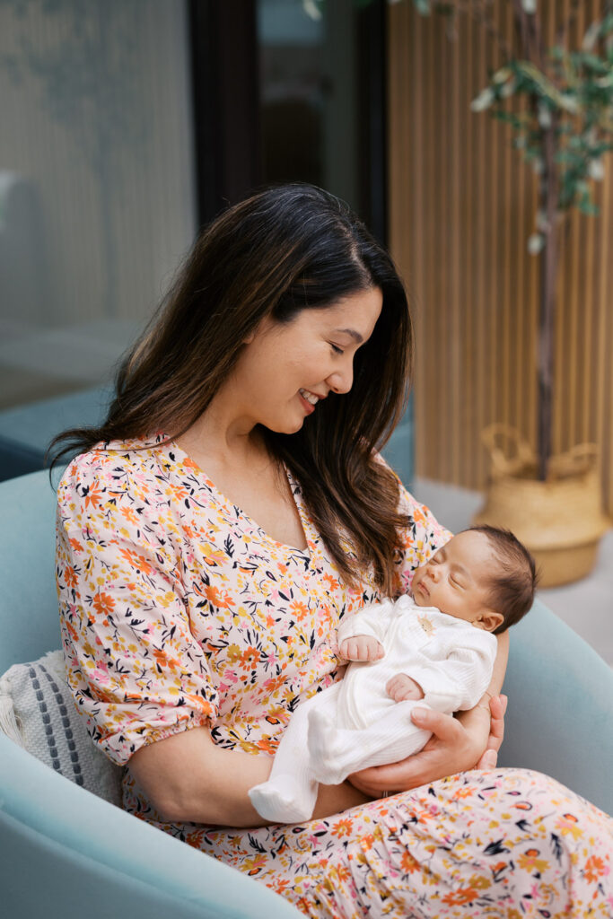 mother in light floral dress holds newborn baby boy while sitting in blue chair during San Mateo newborn photography session