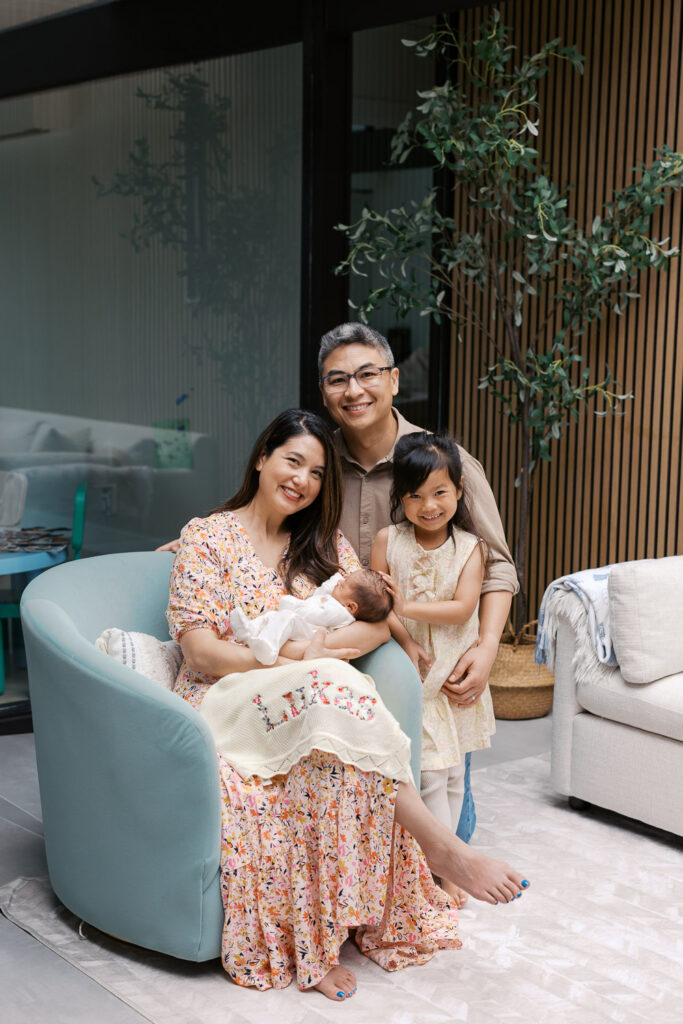 family of four smile at the camera while siting on a blue chair in their home during San Mateo newborn photography session