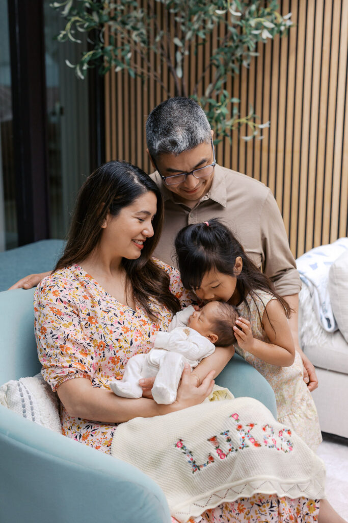 family of four during their San Mateo in home newborn session, toddler leans down to kiss her newborn baby brother