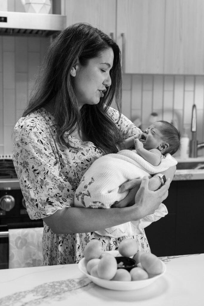Mother holds crying newborn as she shusshs him to calm him down while standing in their kitchen in San Mateo