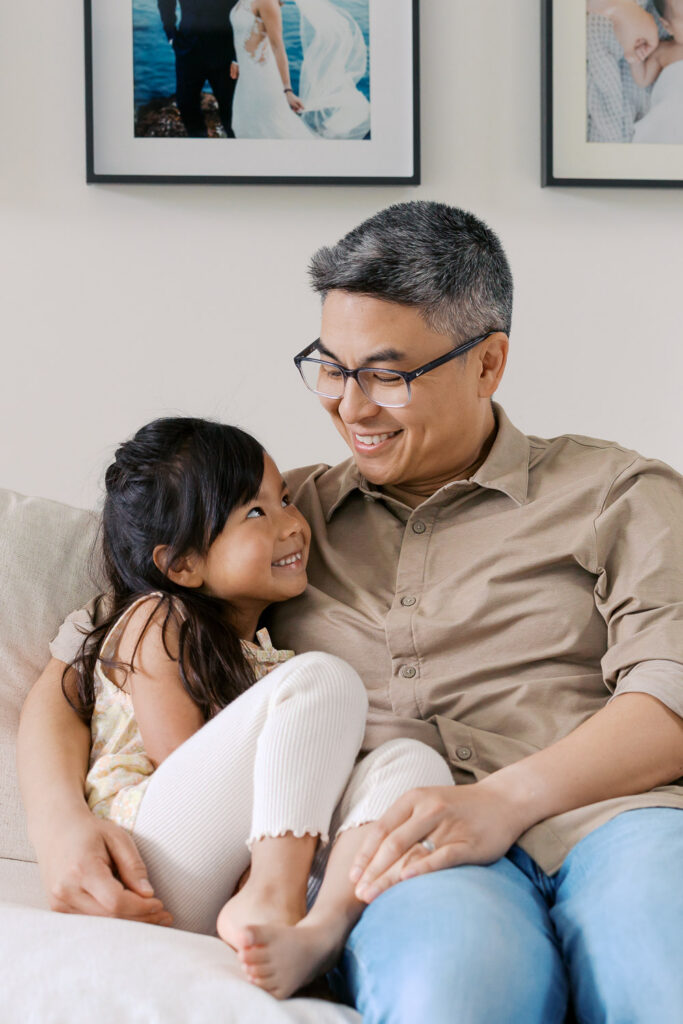 dad and daughter smile at each other while sitting on white couch in San Mateo