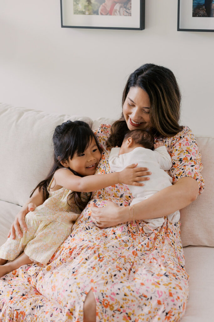 Mom holds newborn baby boy while older sister leans in to show him affection while sitting on a white couch at their home in San Mateo