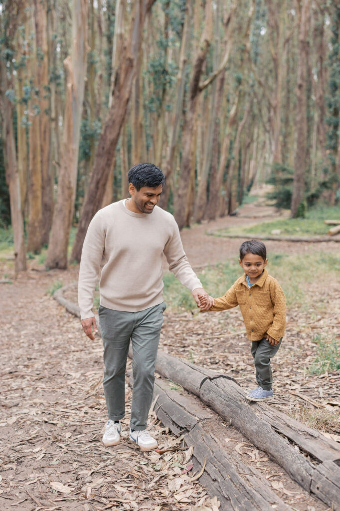 Dad holds toddler's hand while he walks on a tree beam in the Presidio in San Francisco during a photography session