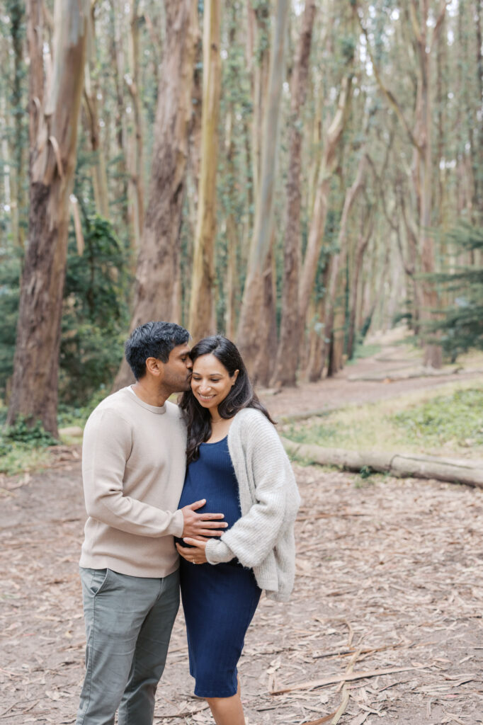 expecting father kisses his pregnant wife on the forehead while they stand next to the Woodline in the Presidio during their maternity session