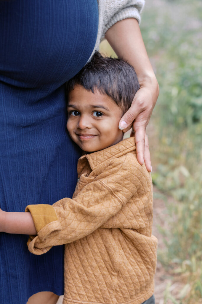 toddler smiles at camera while holding onto his mom during maternity photography session