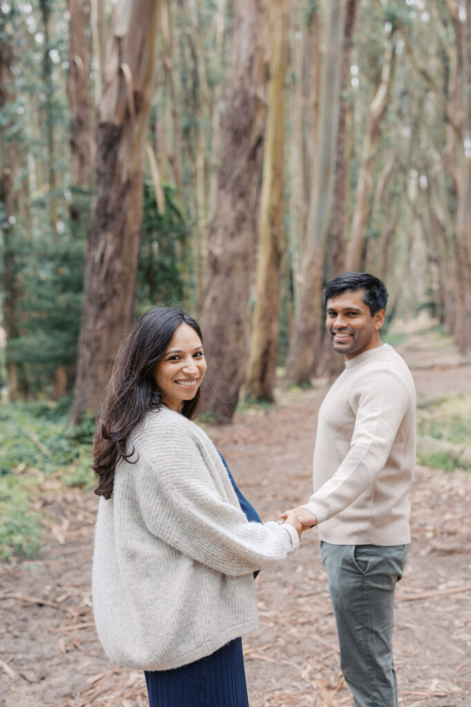 couple hold hands and walk thru the trees in the Presidio during their maternity photography session