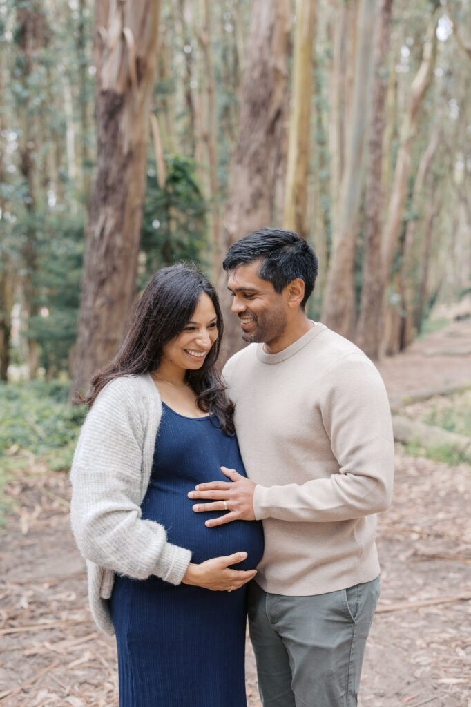expecting couple cuddle in the trees, man gazes at his pregnant wife who laughs at their toddler out of the image