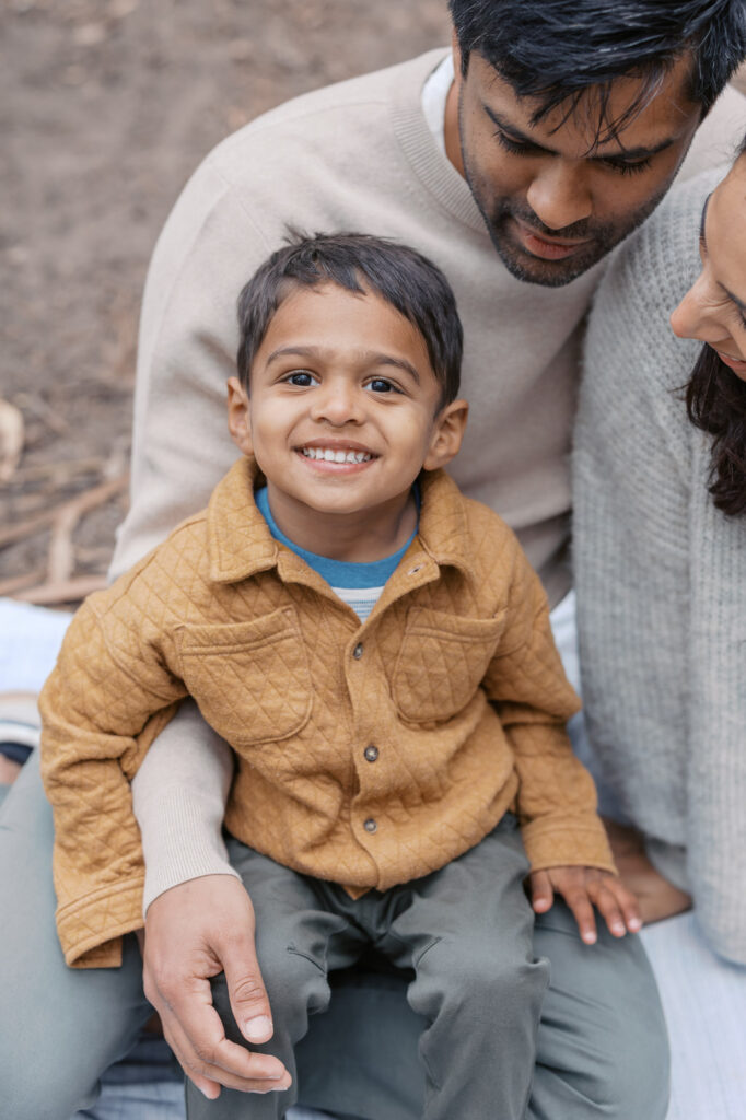 toddler smiles at camera while seated on dad's lap during maternity photography session