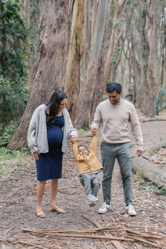 expecting parents swing their toddler while walking along the path at Woodline, Presidio