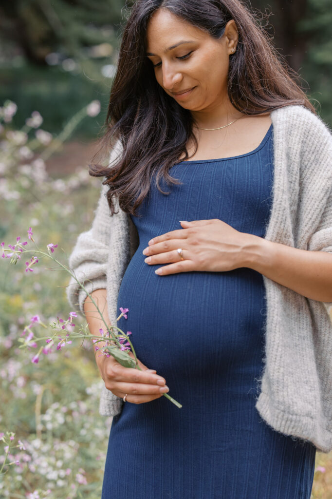 expecting mama in a blue dress holds her pregnant belly and gazes downward while in a field of wildflowers in the Presidio in San Francisco
