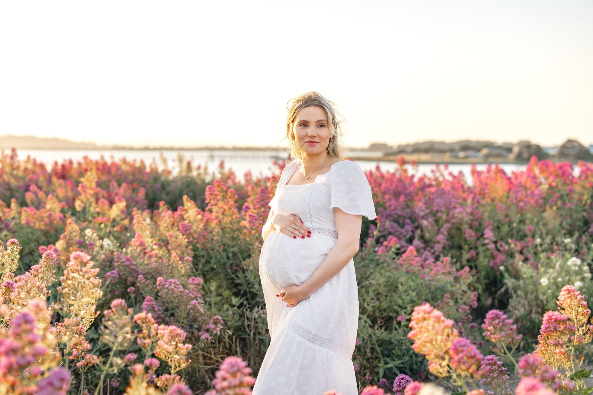 Expecting mother in white dress stands in a field of pink flowers in East Bay during a maternity session
