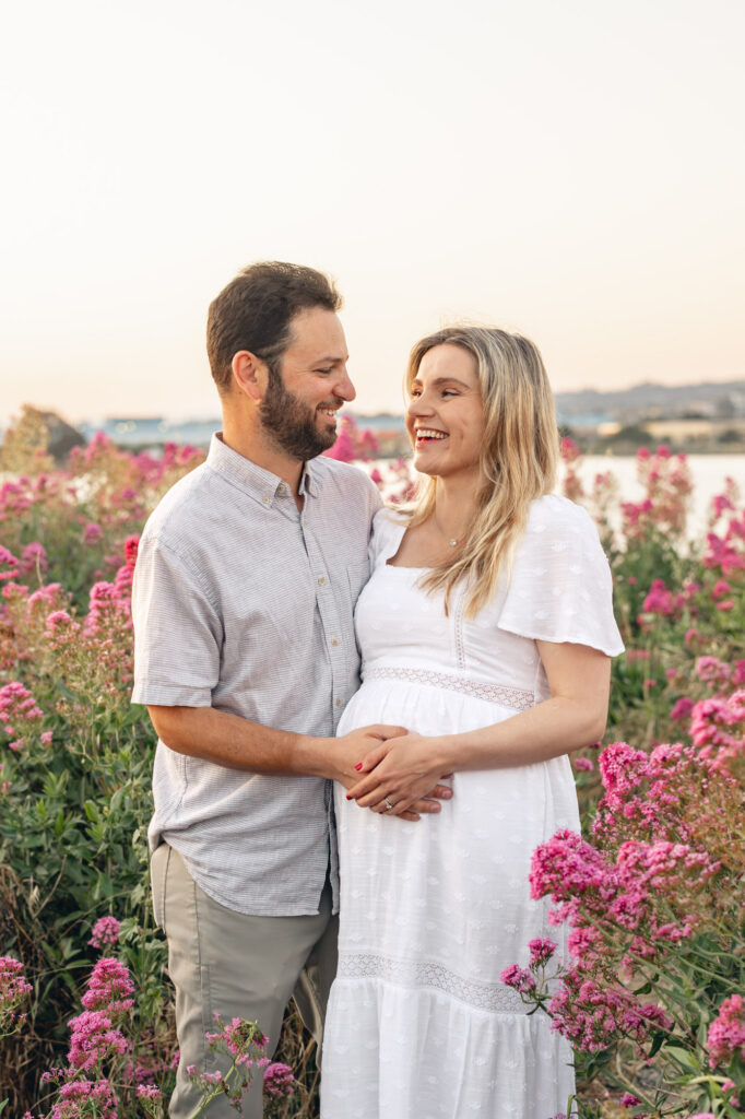 Expecting couple laughs in an East Bay flower field during maternity session in pink flowers