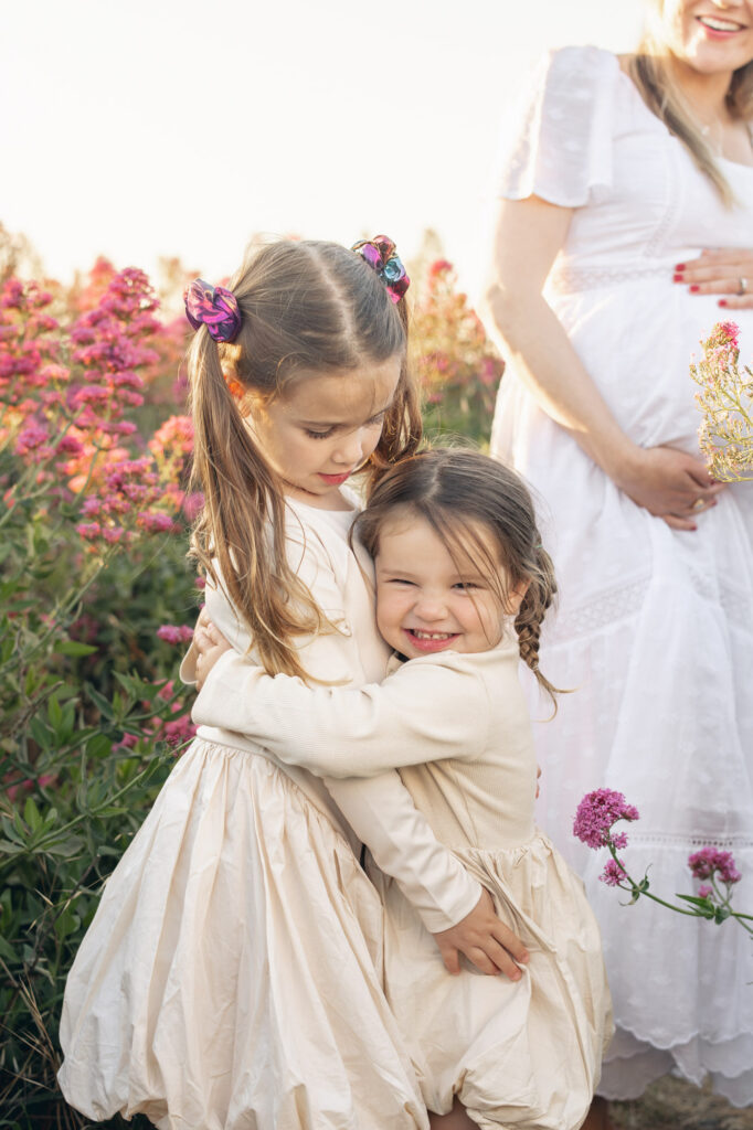 two sisters hug and squeeze each other excitedly while expecting mom looks on laughing