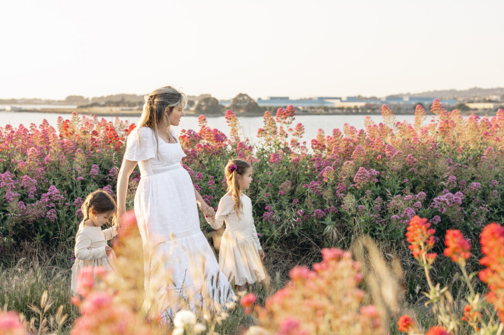 Expecting mother walks thru a pink flower field with her two young daughters during East Bay maternity session