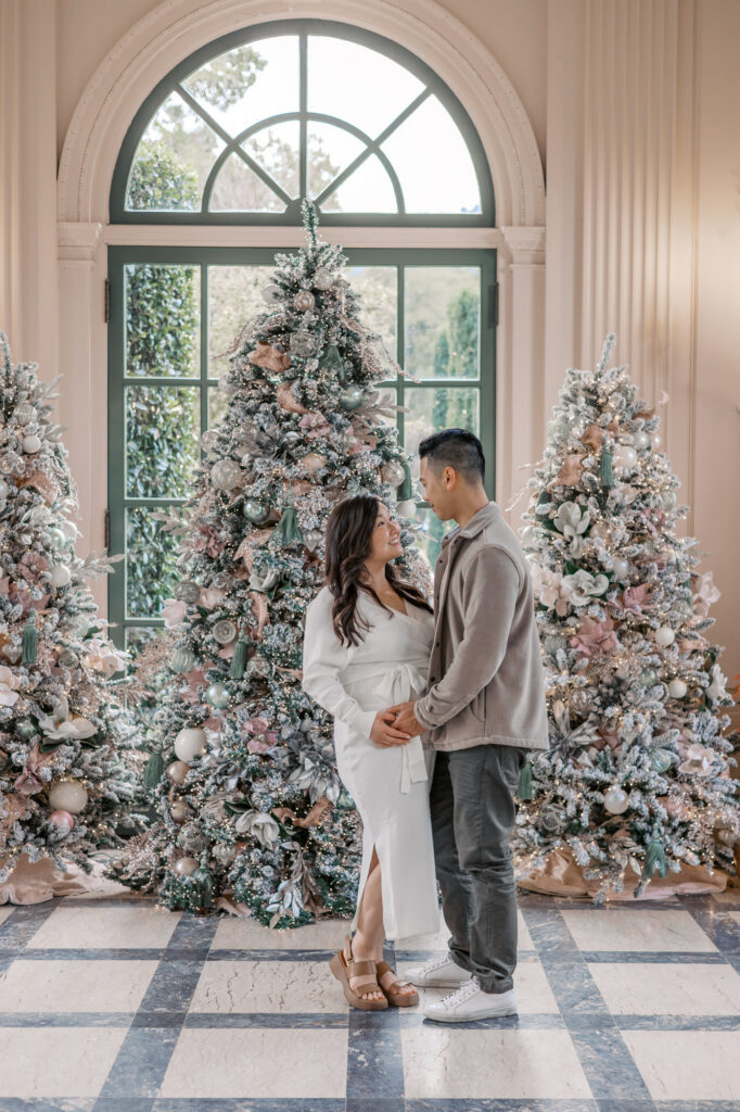 Expecting couple stands in front of Christmas trees at Filoli in the garden house during their Bay Area maternity session