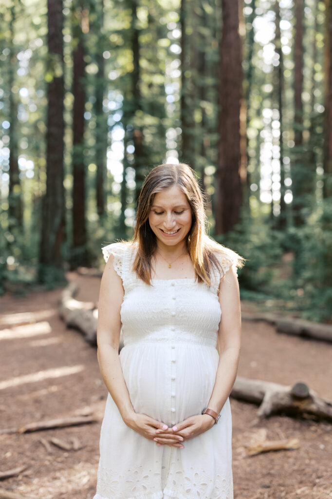 expecting mother in white dress stands in the Oakland redwoods with the light filtering around her during her Bay Area maternity photography session