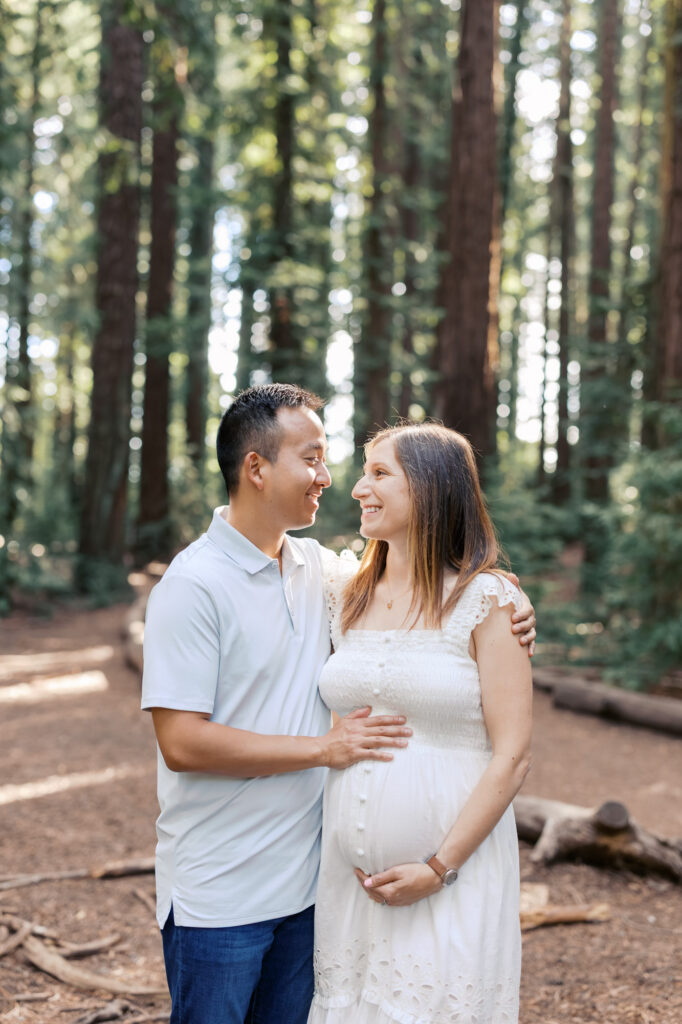 expecting couple stand in the redwoods with light filtering through the trees behind them during their Oakland maternity photography session