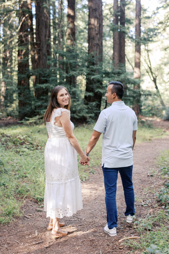 expecting mom glances back at the camera while walking through the redwoods with her husband during their Oakland redwoods maternity photography session