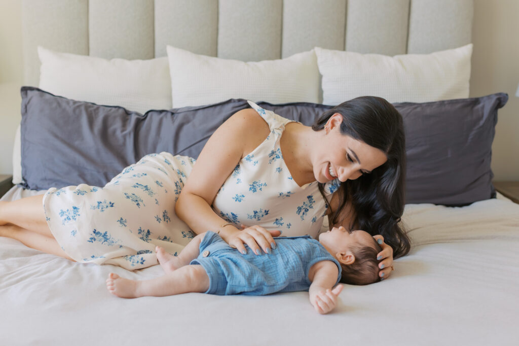 new mom and newborn baby boy at 2 months old lay on a white bed and smile at each other