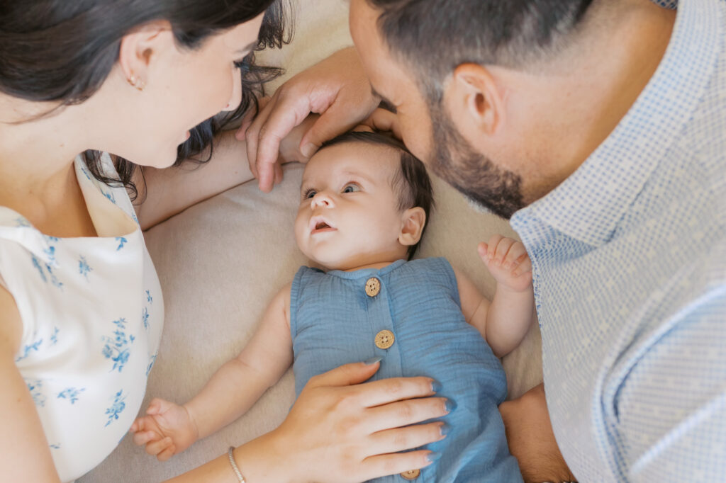 2 month old gazes up at mom and dad as they all lay on a white bed cuddled together