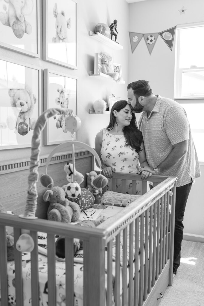 new dad kisses new mom's forehead while they stand next to their 2 month old baby's crib in a football themed nursery in Redwood City, black and white image