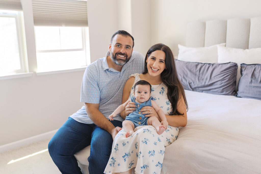 new family of three sits on a white bed holding their 2 month old during Redwood City newborn session