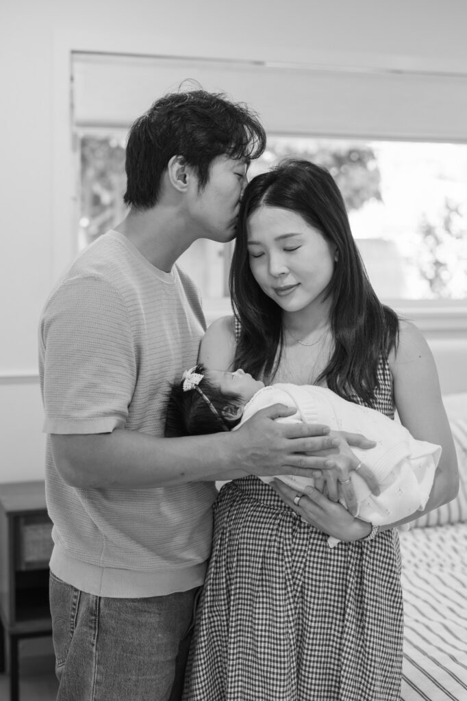 new mom holds newborn in her arms while her husband kisses her forehead during in home newborn photography session in Santa Clara