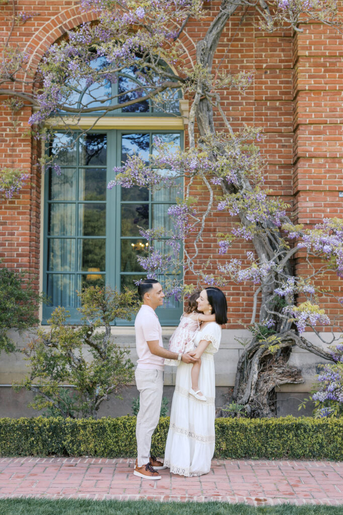 Family of three stands among the wisteria on the brick Filoli estate house during spring